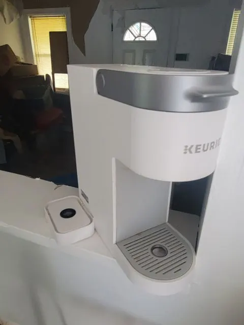 A single-serve coffee maker photographed straight on in a water-damaged kitchen, its brand badge legible